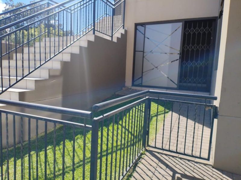 Exterior rear view of the apartment featuring a paved walkway, metal staircase with balustrade, and a secure sliding glass entrance door.
