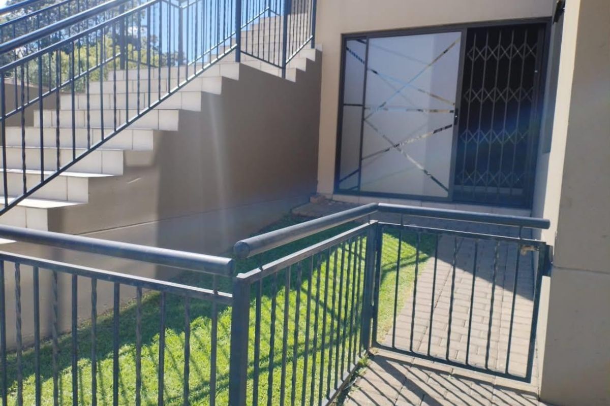Exterior rear view of the apartment featuring a paved walkway, metal staircase with balustrade, and a secure sliding glass entrance door.