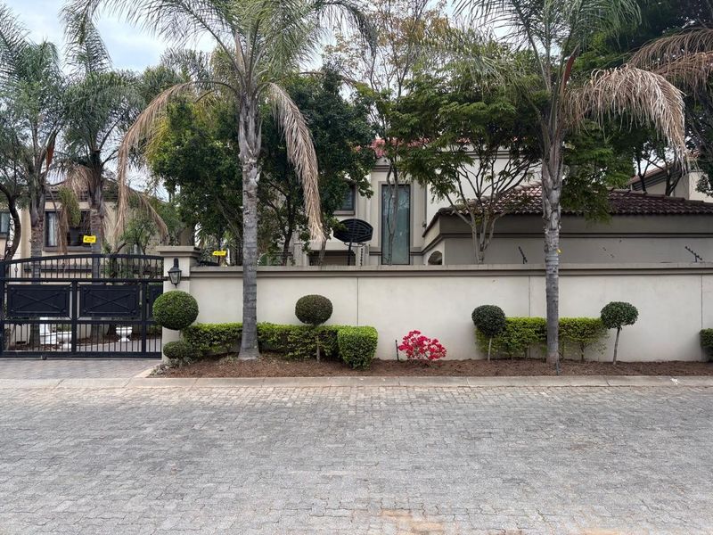 Front view of the property featuring a secure perimeter wall, wrought iron gate, paved driveway, and established garden landscaping.