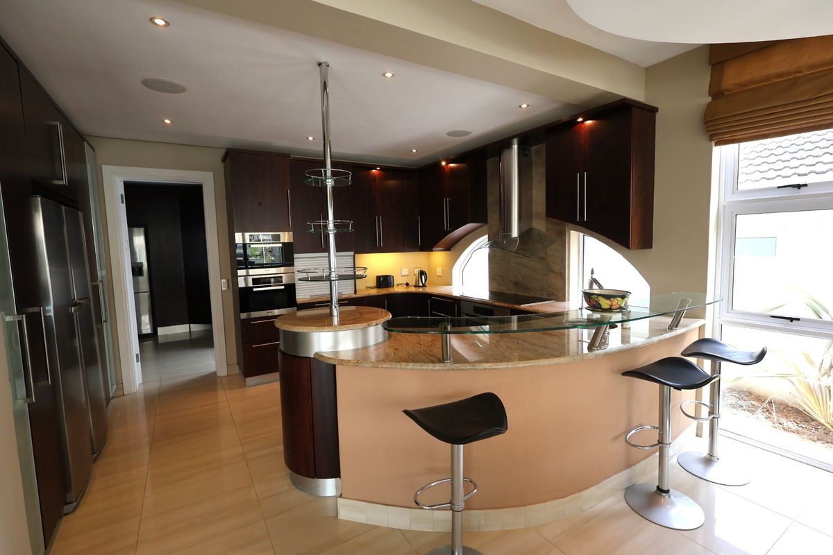 Modern kitchen featuring dark wood cabinetry, granite countertops, a curved glass breakfast bar, built-in appliances, and recessed lighting with tiled flooring.