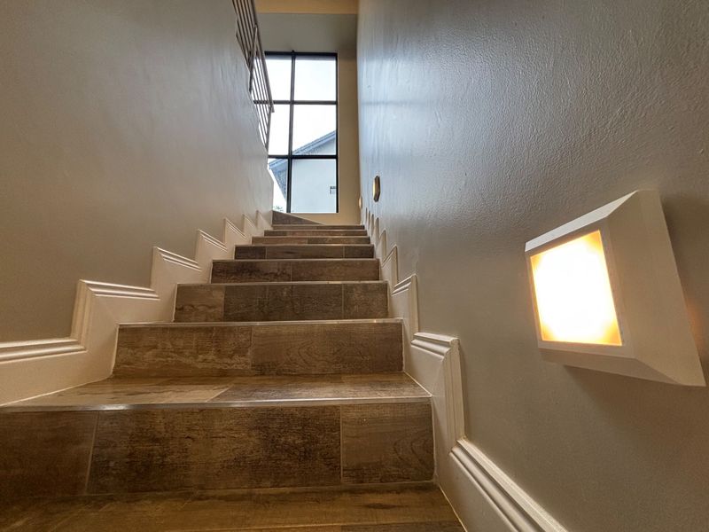 A well-lit staircase featuring modern wood-look tiled steps, clean wall finishes, and integrated wall lighting leading to a large window.