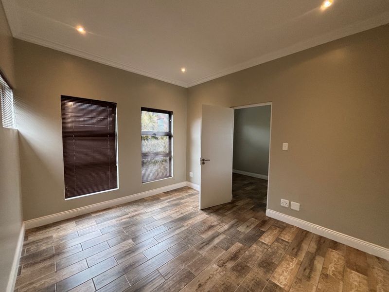 A bright, empty bedroom featuring modern wood-look tile flooring, recessed downlighting, and venetian blinds, offering a clean and neutral space ready for furnishing.