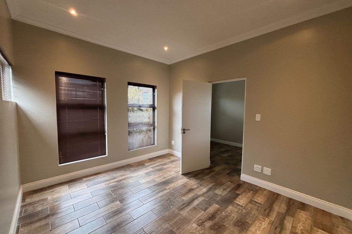 A bright, empty bedroom featuring modern wood-look tile flooring, recessed downlighting, and venetian blinds, offering a clean and neutral space ready for furnishing.