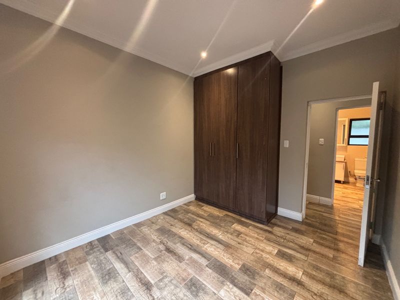 A clean, empty bedroom featuring wood-look tile flooring, built-in dark wood cupboards, and recessed lighting, with an adjoining bathroom visible through the doorway.