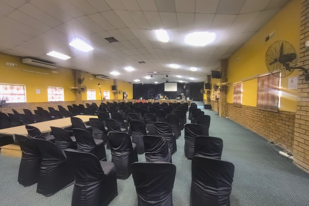 A large commercial event hall or clubhouse featuring a stage, suspended ceiling, wall-mounted fans, and air conditioning units, suitable for gatherings or presentations.