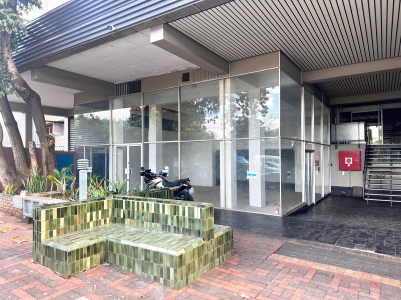 Commercial building entrance featuring a glass facade, metal ceiling soffit, and a tiled outdoor bench area with adjacent metal staircase.