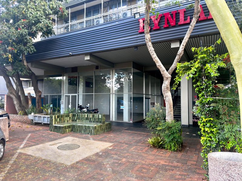 Front facade of a commercial building featuring a glass curtain wall, brick paved entrance area, and a built-in tiled bench.