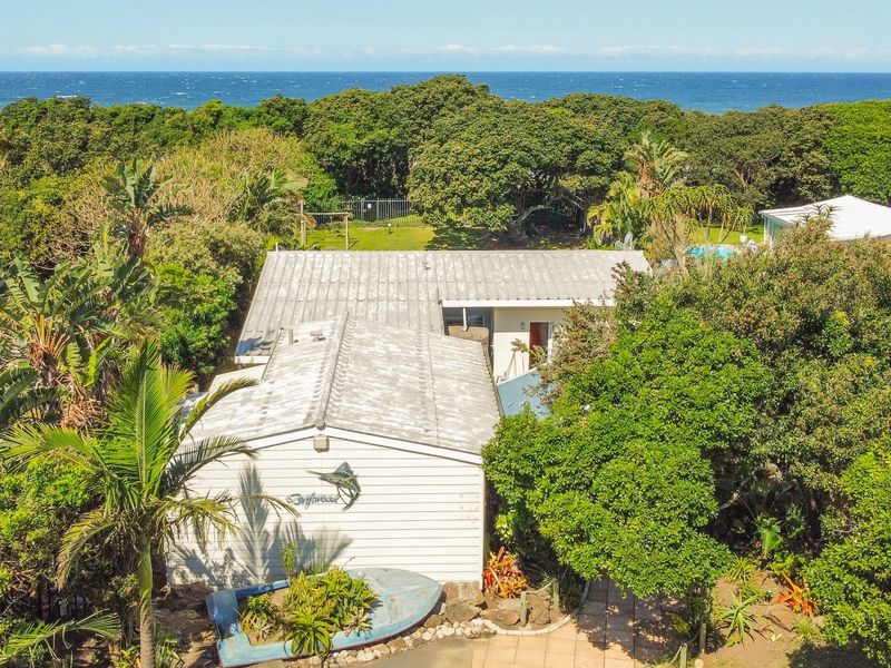Aerial view of a coastal property featuring a metal roof, weatherboard exterior, and lush surrounding vegetation with ocean views in the background.