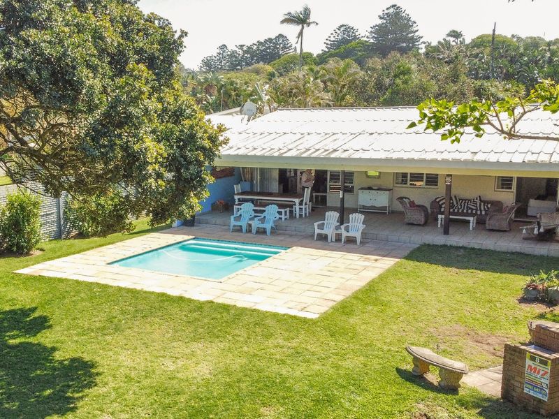 Rear exterior view featuring a swimming pool, covered patio area, and a built-in brick braai set within a spacious garden.