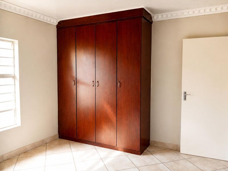 A bright, empty bedroom featuring a large, dark wood built-in wardrobe, tiled flooring, and decorative ceiling cornicing.
