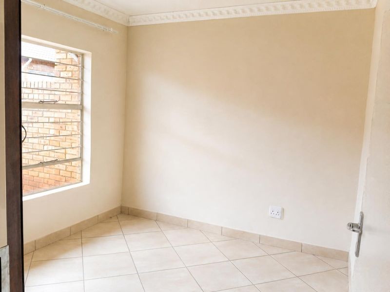 A clean, empty bedroom featuring neutral tiled flooring, cornice detailing, and a window fitted with security bars.