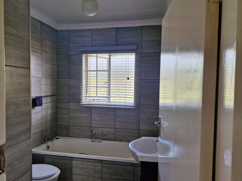 A clean, modern-tiled bathroom featuring a bathtub, pedestal basin, and toilet, finished with neutral grey wall tiles and a functional window.