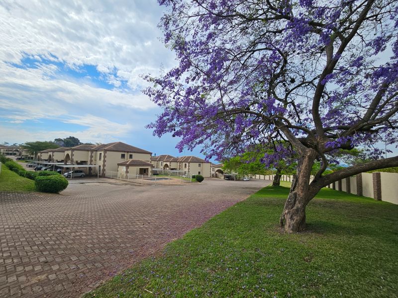 A wide-angle view of the apartment complex exterior, featuring paved driveways, established gardens with a large jacaranda tree, and covered parking areas.