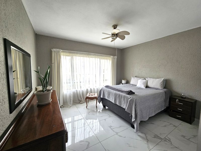 A well-lit bedroom featuring large windows, a ceiling fan, and modern marble-look tiled flooring.