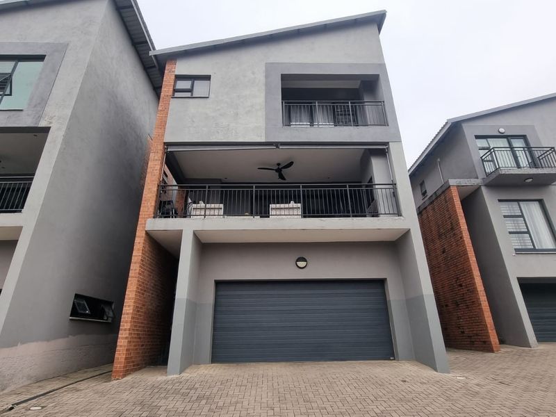 Modern multi-story townhouse featuring a double garage, paved driveway, and a spacious balcony with metal railings and a ceiling fan.