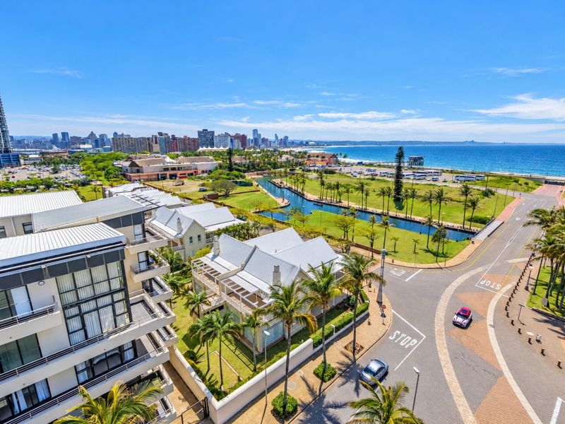 An elevated aerial view showcasing the surrounding coastal neighborhood, including nearby residential buildings, landscaped parklands, a waterway, and proximity to the beach.