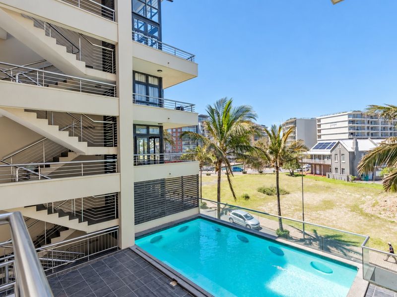 An elevated view of a modern apartment complex featuring a swimming pool, tiled deck, and external staircases with stainless steel railings.