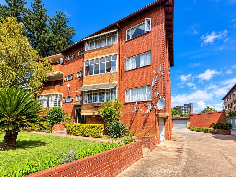 A multi-story brick apartment building featuring casement windows, a concrete driveway, and established landscaping with a brick retaining wall.