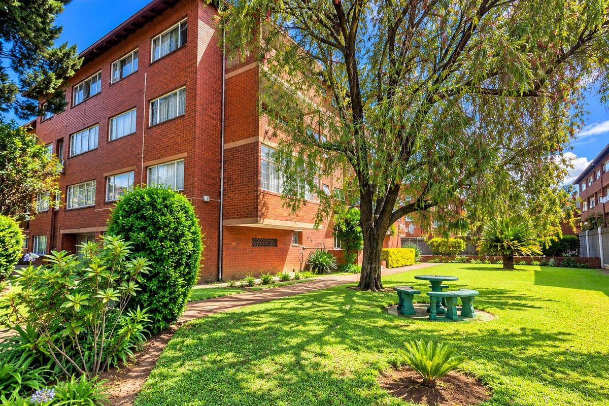 A view of the rear exterior of a multi-story brick apartment building featuring a well-maintained garden, mature trees, and a paved walkway.