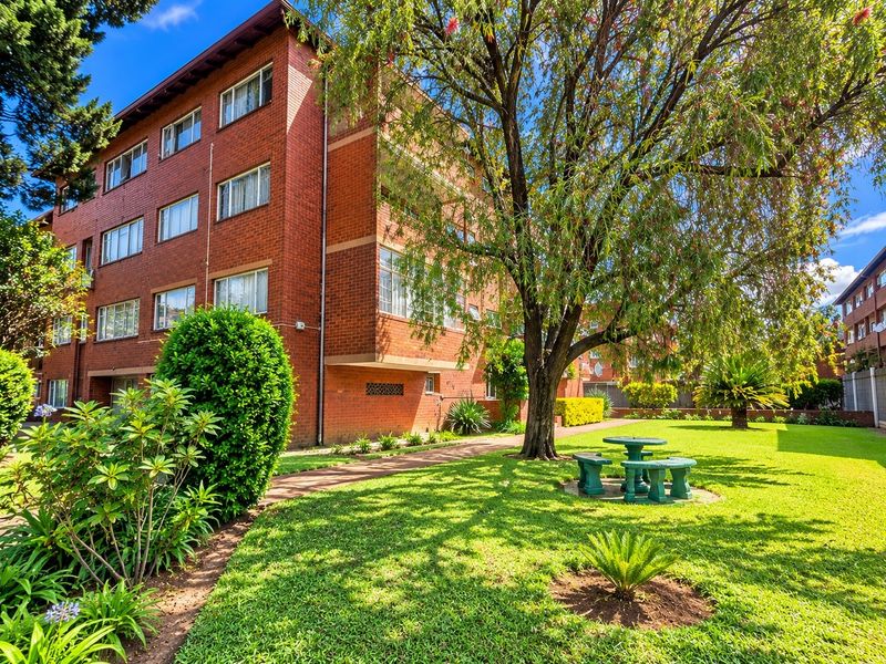 A view of the rear exterior of a multi-story brick apartment building featuring a well-maintained garden, mature trees, and a paved walkway.
