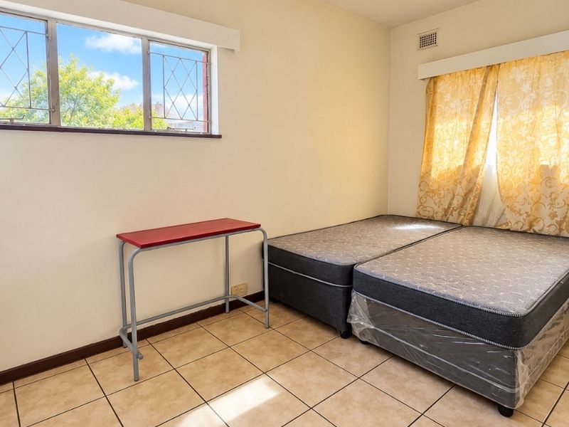 A functional bedroom featuring tiled flooring, a ceiling pendant light, and metal-framed windows, offering a neutral canvas for potential interior updates.