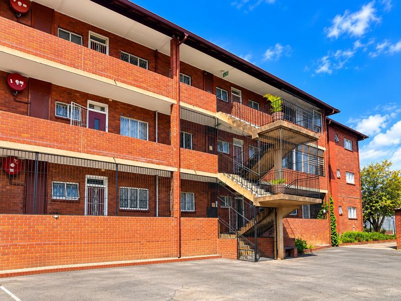 Exterior view of a multi-story brick apartment building featuring external concrete staircases, security grilles on windows, and an asphalt parking area.