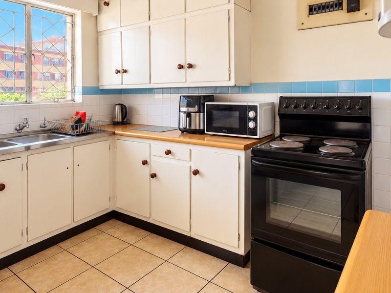A functional kitchen featuring built-in cabinetry, a stainless steel sink, tiled splashback, and a freestanding electric stove with a visible electrical distribution board.