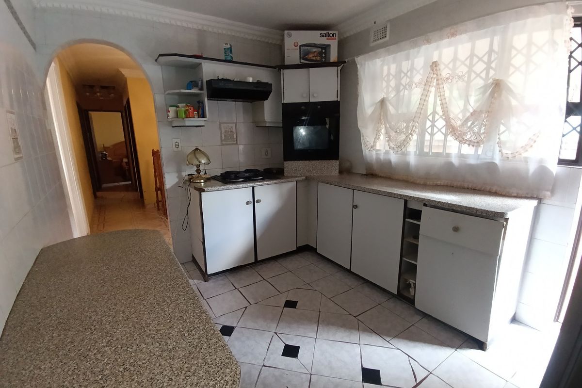 A dated kitchen featuring built-in cabinetry, laminate countertops, integrated oven and hob, and tiled flooring with an adjacent hallway archway.