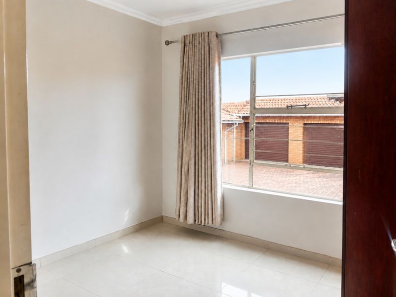 A bright, empty bedroom featuring neutral tiled flooring, coving, and a large window with a curtain rod, offering a view of the complex exterior.