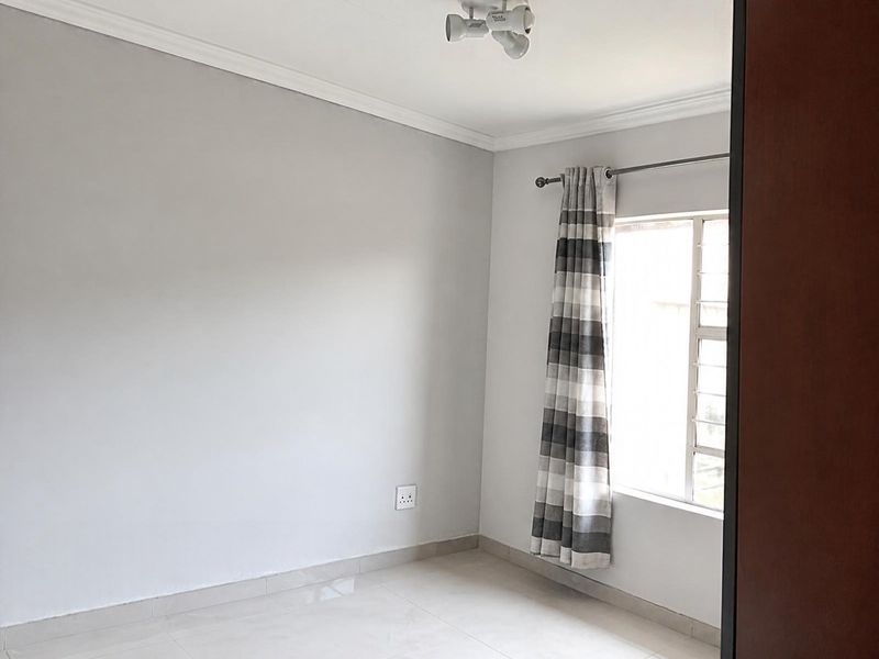 A clean, neutral-toned bedroom featuring tiled flooring, a window with security bars, and modern ceiling lighting, offering a blank canvas for personalization.