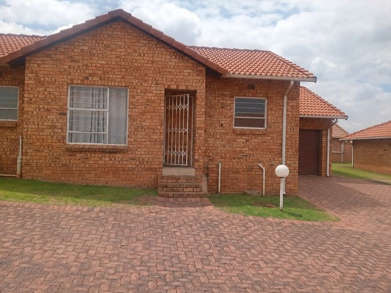 Front elevation of a face brick townhouse featuring a tiled roof, paved driveway, and secure entrance with security gate.
