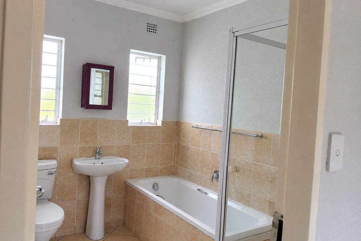 A functional bathroom featuring a pedestal basin, toilet, and a bathtub with a shower screen, finished with neutral wall and floor tiling.