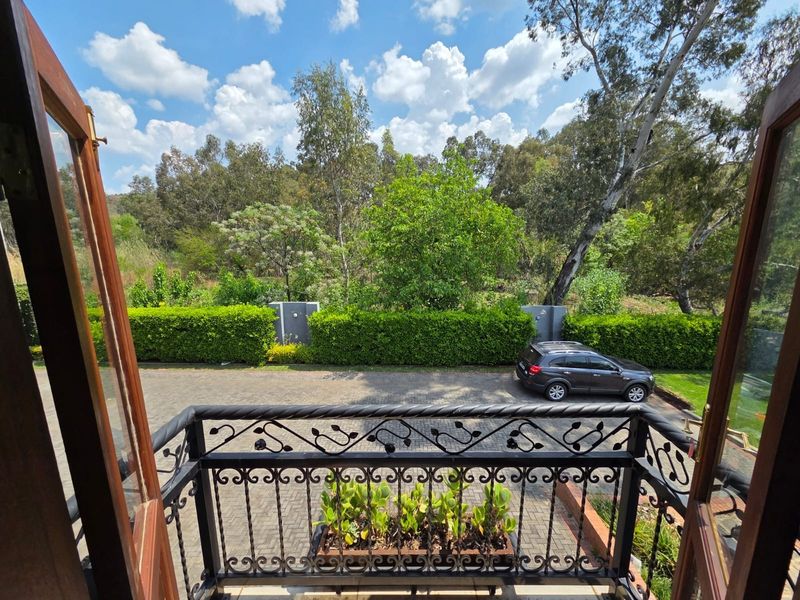 A view from a balcony featuring a decorative wrought iron balustrade overlooking a paved driveway and lush garden surroundings.