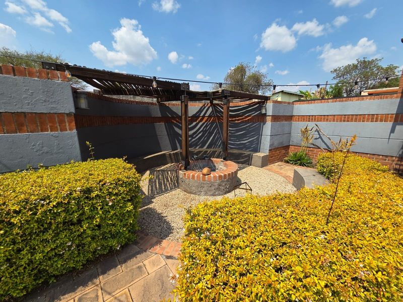 An outdoor entertainment area featuring a central brick fire pit, timber pergola, and landscaped garden beds enclosed by a perimeter wall.