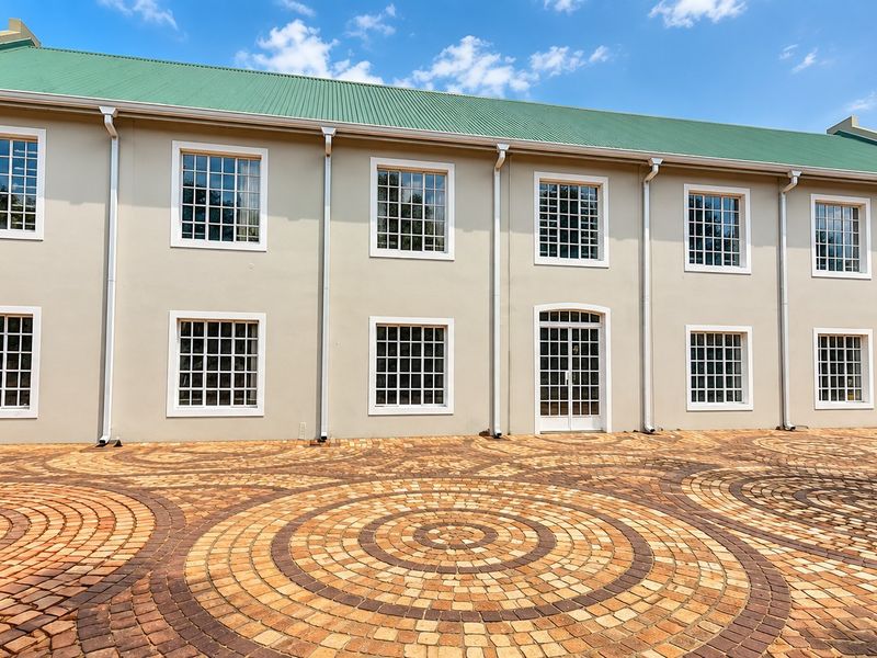 A well-maintained rear exterior view of a retail building featuring a green corrugated roof, grid-pattern windows, and a decorative circular paver courtyard.