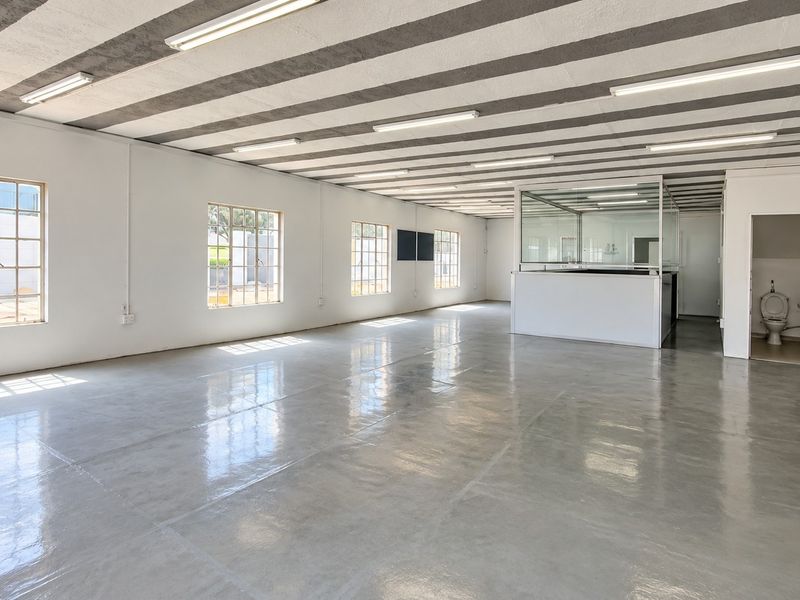 Spacious open-plan retail or office area featuring polished concrete floors, exposed beam ceilings, ample natural light from grid windows, and a private partitioned office space.