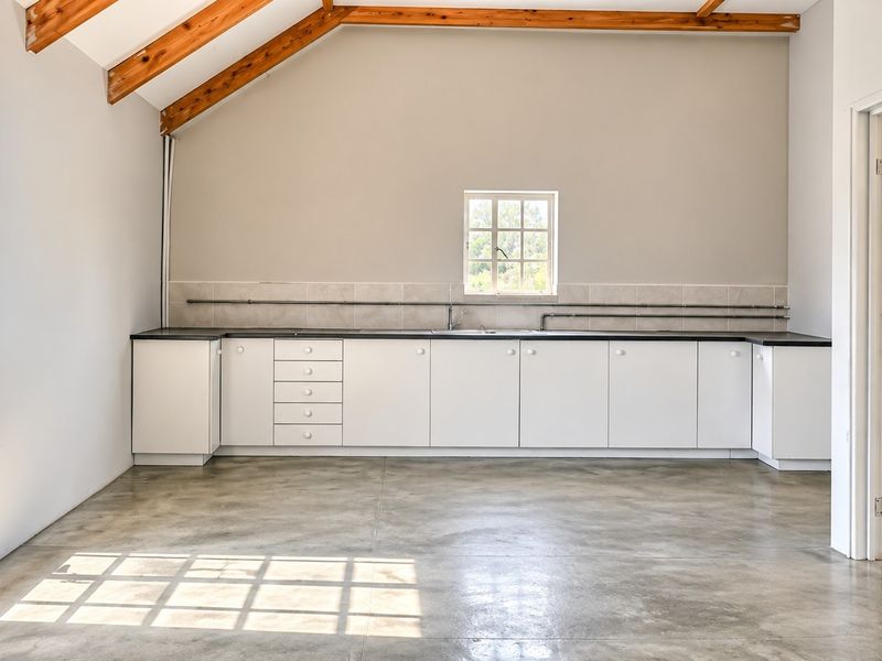 A bright, empty kitchen space featuring exposed timber beams, concrete flooring, white cabinetry, and an arched window, offering a clean, modern industrial aesthetic.