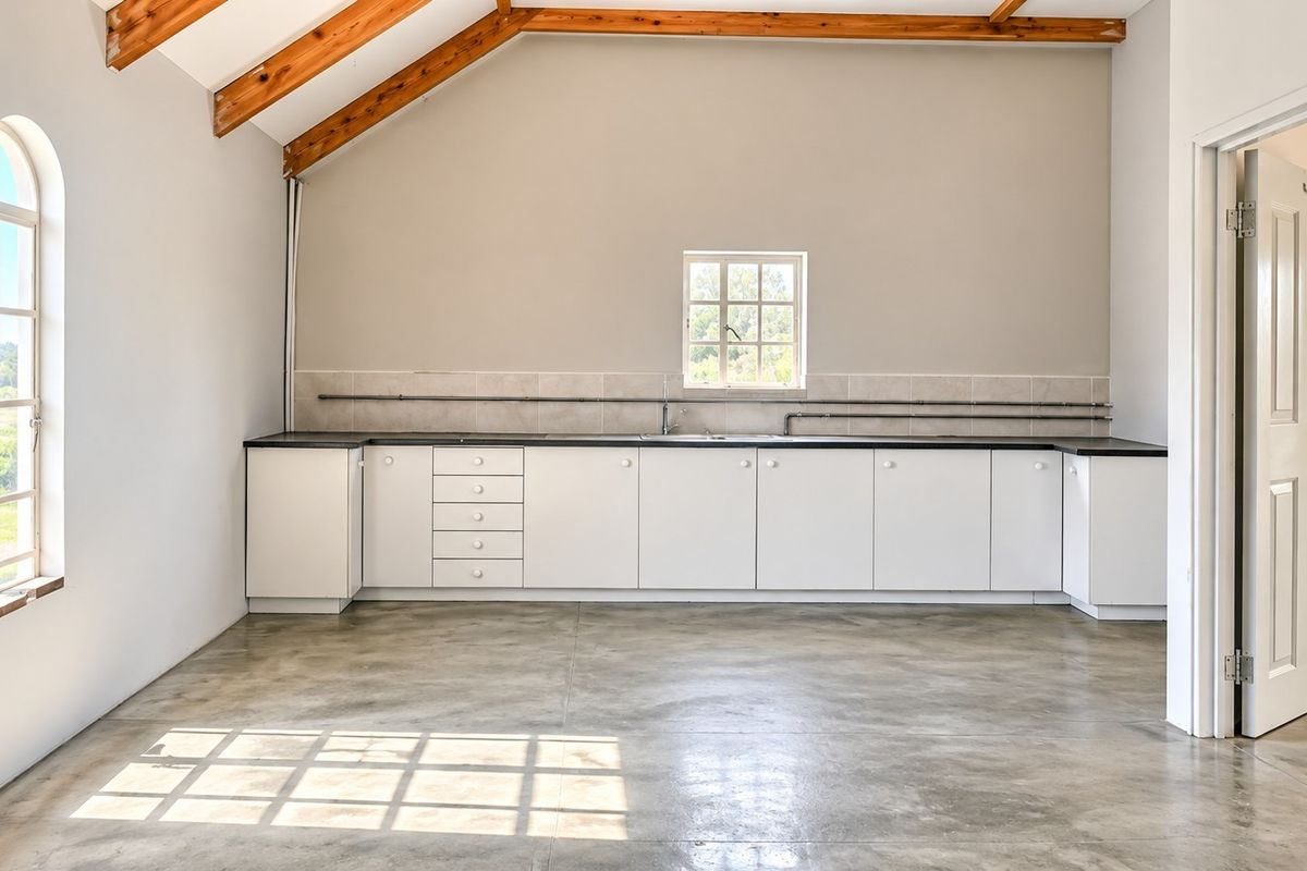 A bright, empty kitchen space featuring exposed timber beams, concrete flooring, white cabinetry, and an arched window, offering a clean, modern industrial aesthetic.