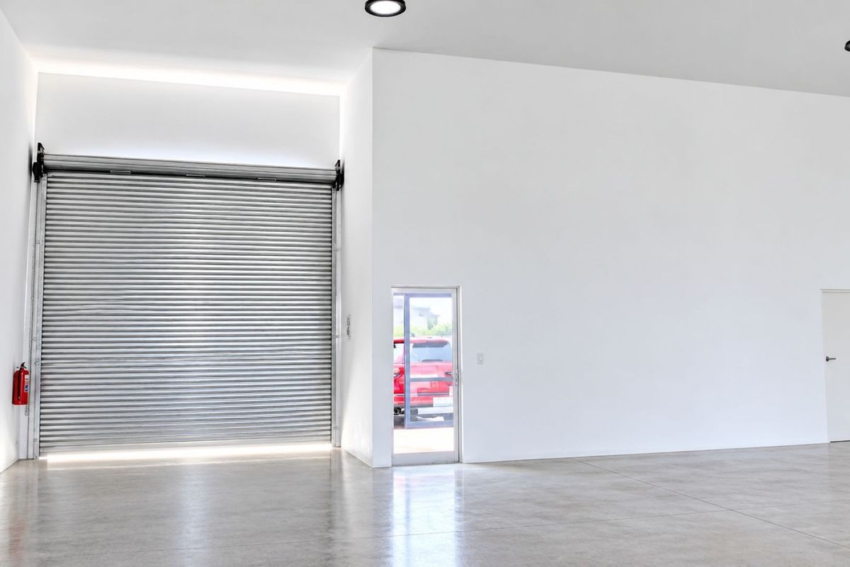 Spacious, modern commercial garage featuring a high-clearance roller shutter door, polished concrete flooring, and clean, minimalist white walls with recessed lighting.