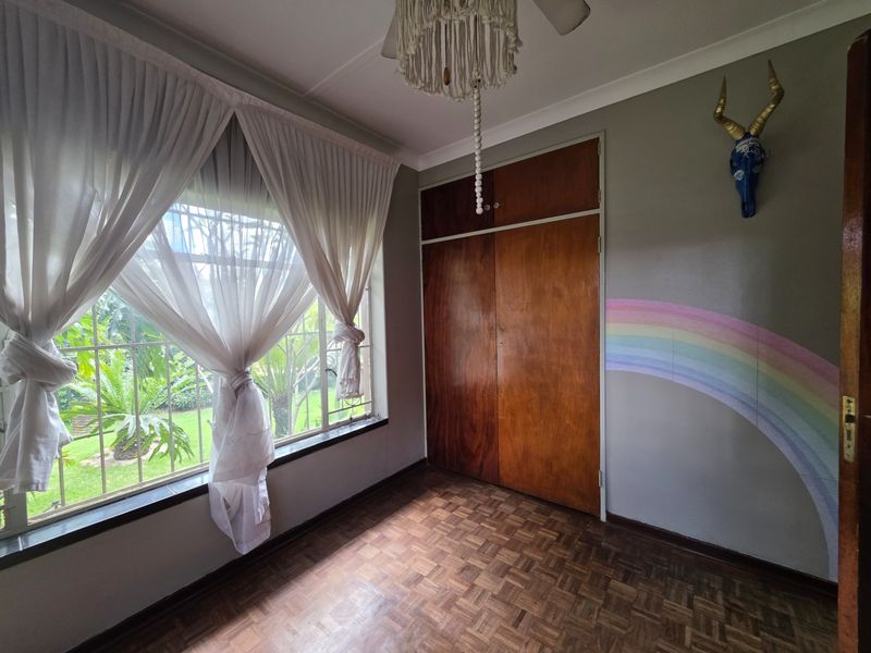 A bright bedroom featuring classic parquet flooring, built-in wooden cupboards, and large windows with security bars overlooking the garden.
