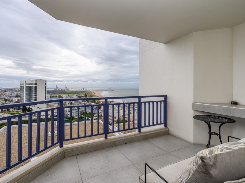 A spacious balcony featuring tiled flooring, a built-in stone counter, and a metal railing, offering expansive coastal and city views.