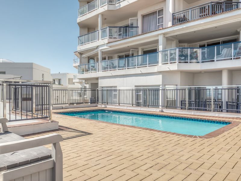 A well-maintained communal swimming pool area featuring paved decking and metal safety balustrades, set against a multi-story apartment building with private balconies.