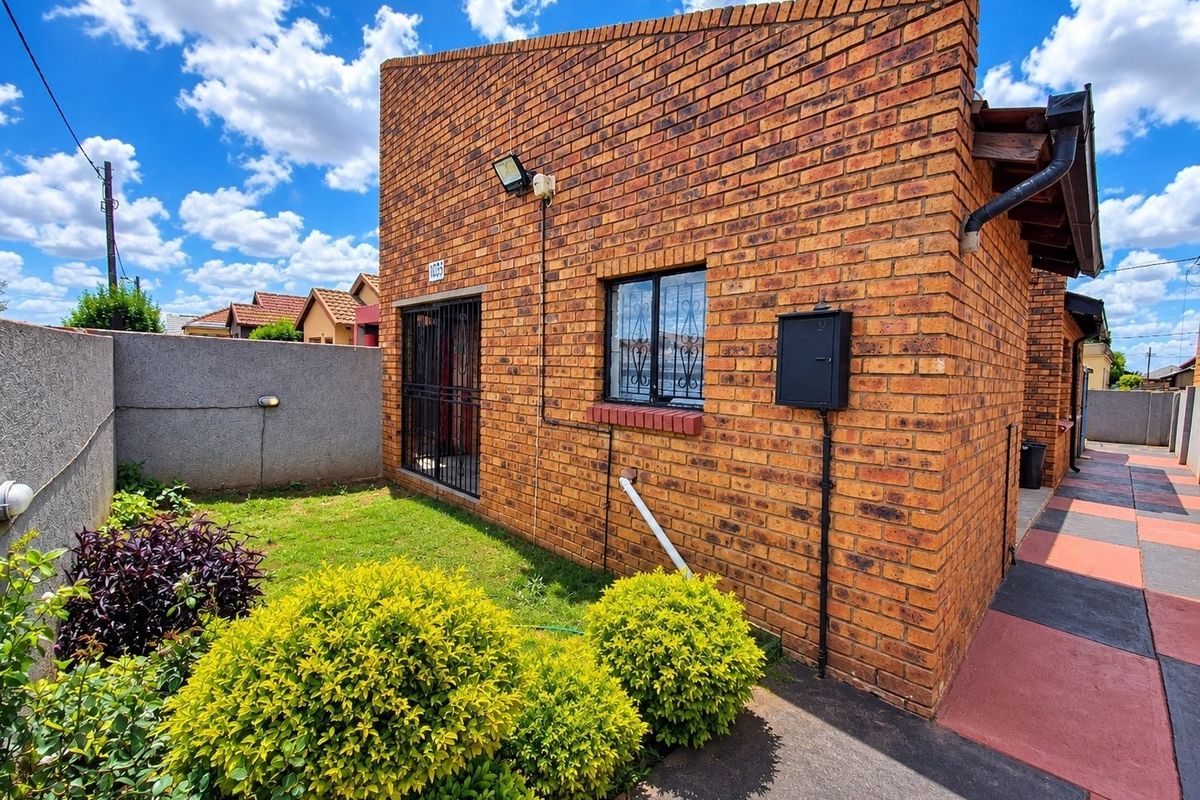 A rear exterior view of a face brick house featuring a small garden, paved walkway, and security fixtures on the windows and doors.