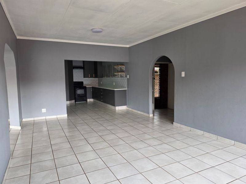 An open-plan living and kitchen area featuring neutral grey walls, tiled flooring, and a functional kitchen with built-in cabinetry and subway tile backsplash.