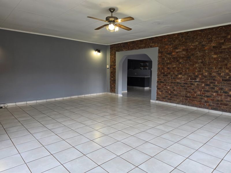 Spacious living area featuring tiled flooring, an exposed brick feature wall, and a ceiling fan, with an arched entryway leading to the kitchen.