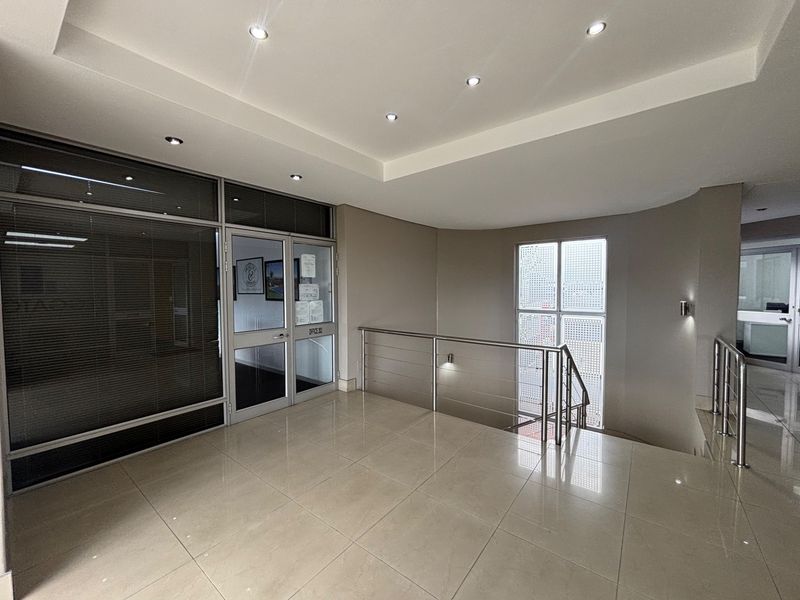 A bright and modern office building hallway featuring tiled floors, recessed lighting, and a stainless steel balustrade overlooking the stairwell.