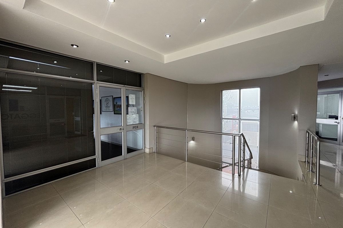 A bright and modern office building hallway featuring tiled floors, recessed lighting, and a stainless steel balustrade overlooking the stairwell.
