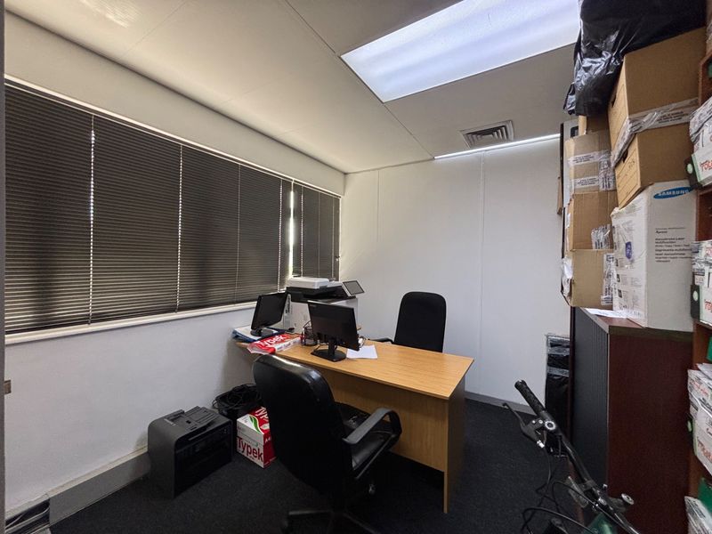 A functional office space featuring carpet flooring, a drop ceiling with recessed lighting, air conditioning ventilation, and large windows with venetian blinds.