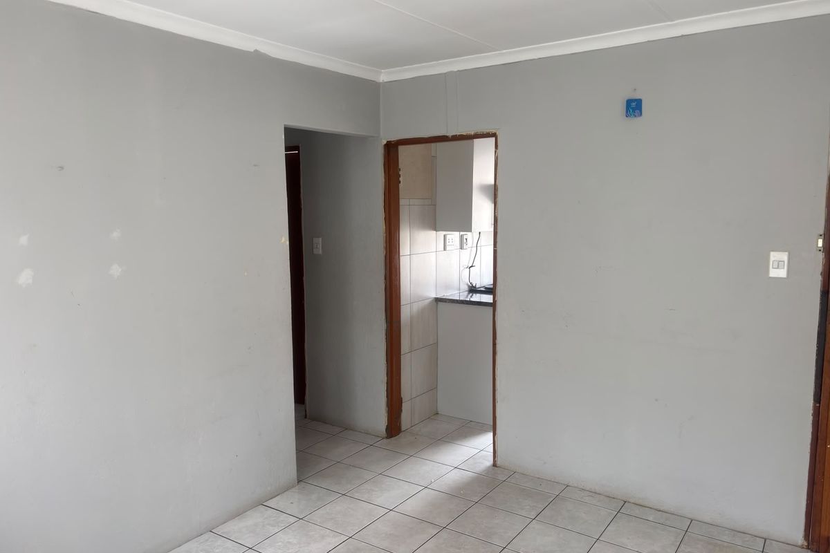 An empty living room featuring neutral grey walls, ceramic floor tiles, and a doorway leading to a kitchen area.