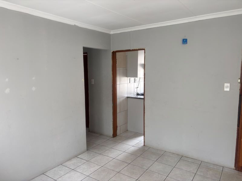 An empty living room featuring neutral grey walls, ceramic floor tiles, and a doorway leading to a kitchen area.